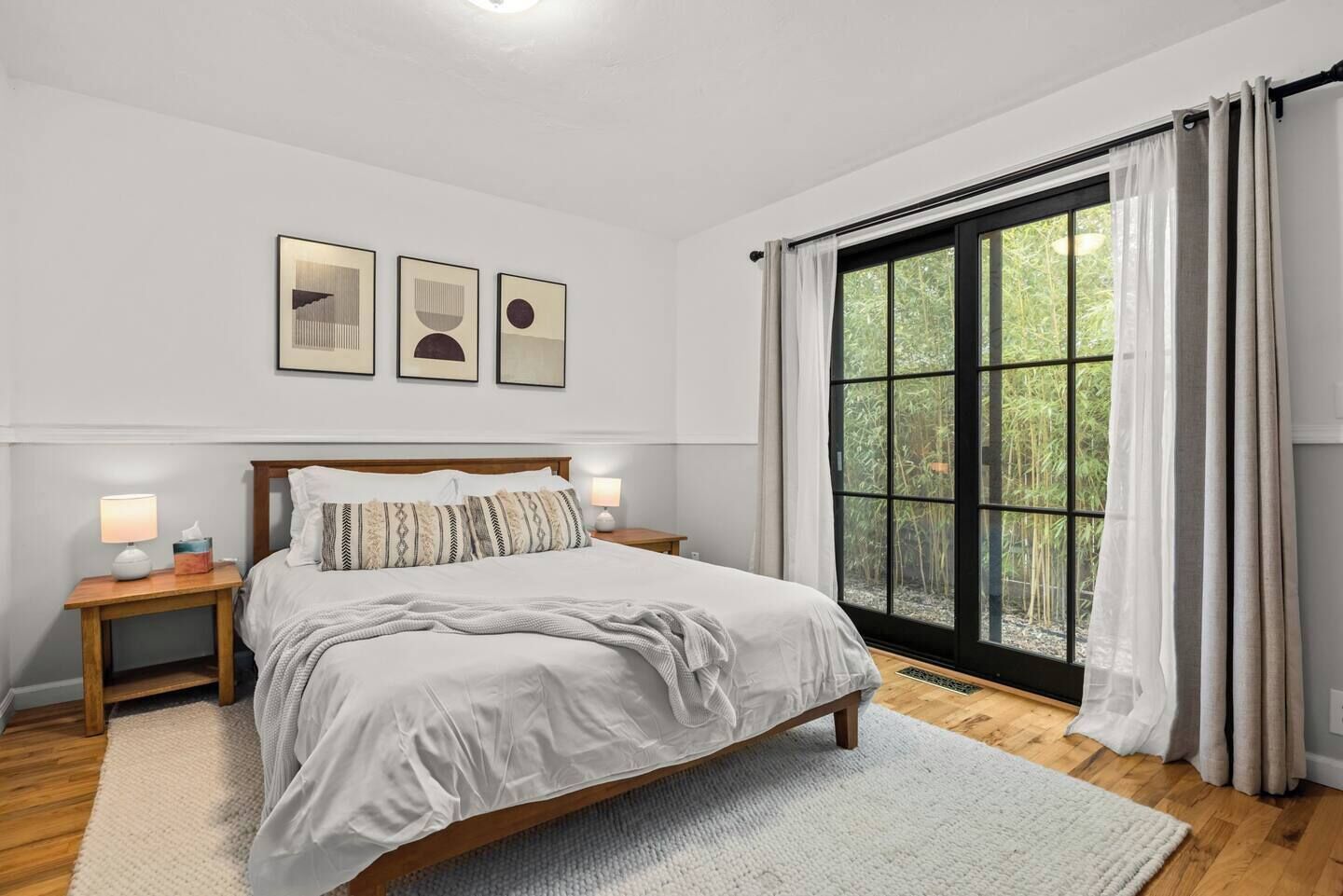 Una camera da letto con un letto grande, una testiera in legno, comodini con lampade, una finestra con tende e opere d'arte incorniciate al muro.