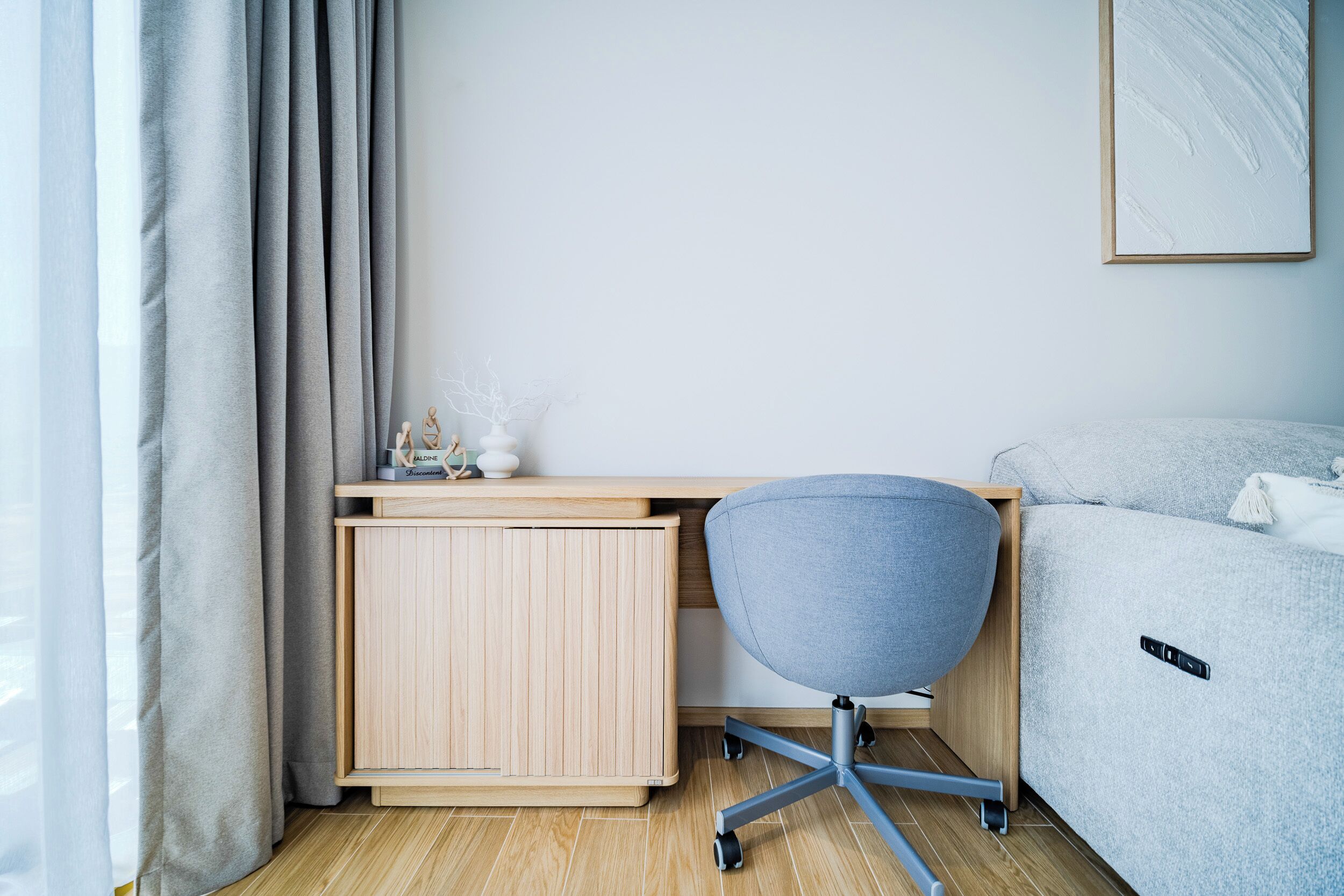 Un dormitorio moderno con un escritorio de madera, una silla de oficina gris, un sofá gris y una mesita de noche blanca.