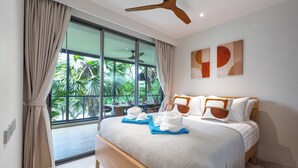 A modern bedroom with a large bed, a ceiling fan, and a view of lush greenery outside. - Beachside Residences by NLA (Choeng Thale)