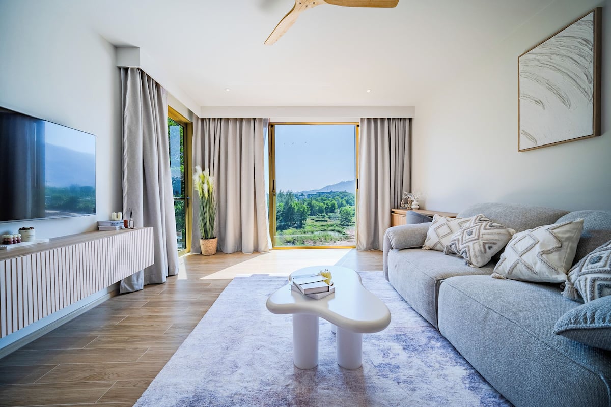 A modern living room with a large TV, a sofa, a coffee table, and a view of a green landscape through a large window.
