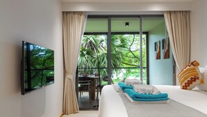 A modern hotel room with a large bed, a flat-screen TV mounted on the wall, a dining area with a table and chairs, and a view of lush greenery through the window. - Beachside Residences by NLA (Choeng Thale)