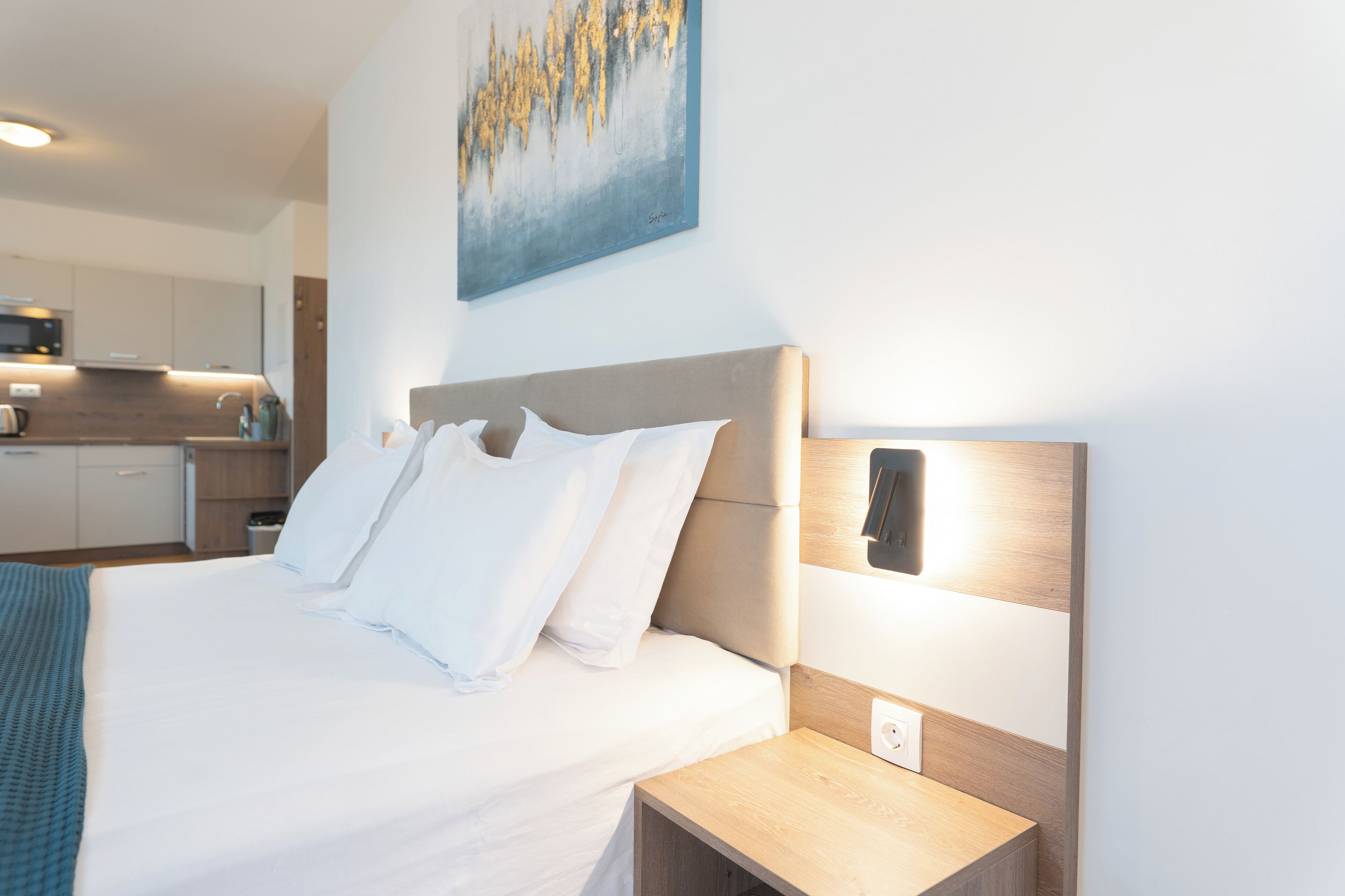 A modern hotel room with a large bed, a bedside table, a wall-mounted light, and a kitchen area in the background.