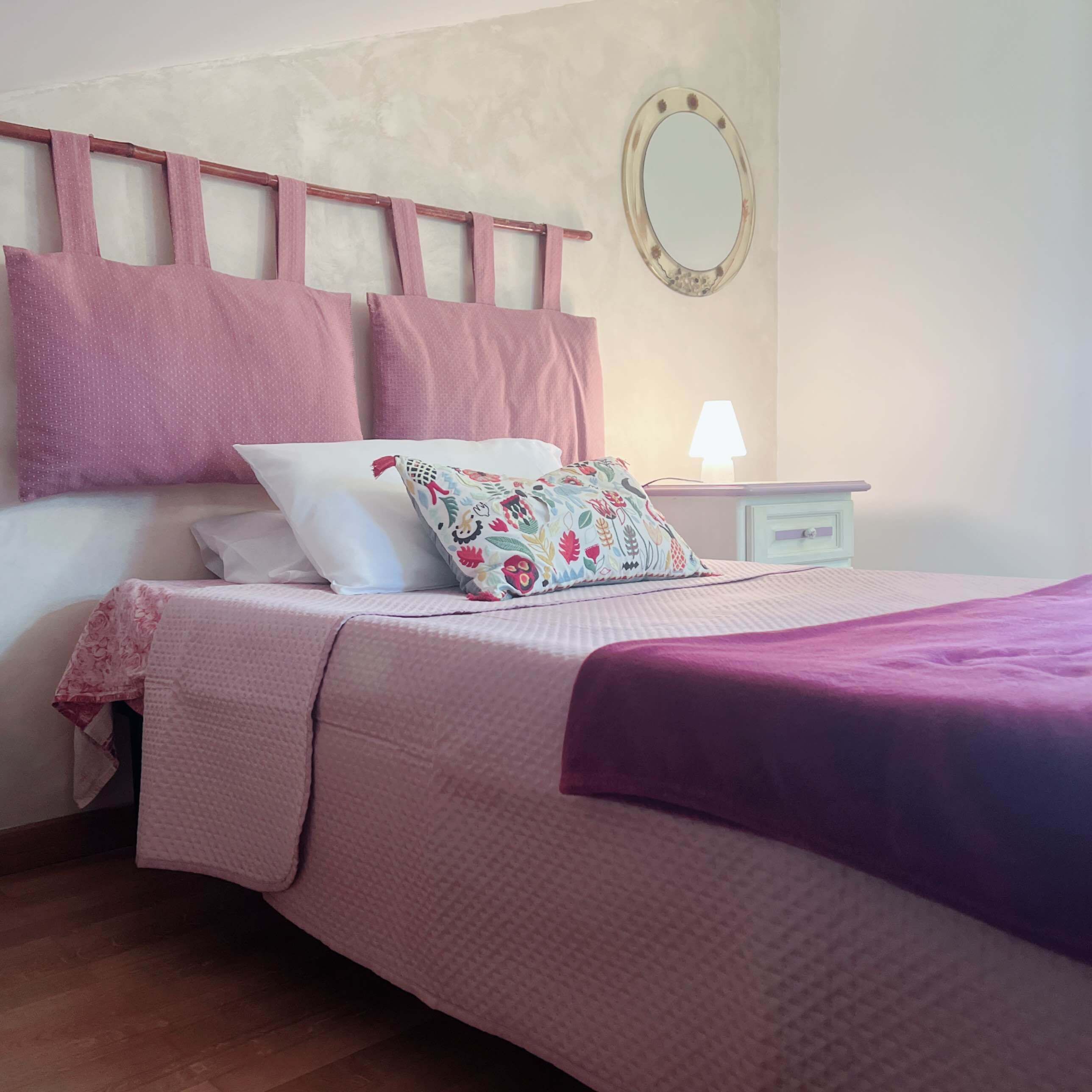 Una cama bien hecha con colcha rosa y blanca, una manta morada y una almohada con motivos florales. Hay una mesita de noche pequeña con una lámpara y un espejo en la pared sobre la cama.