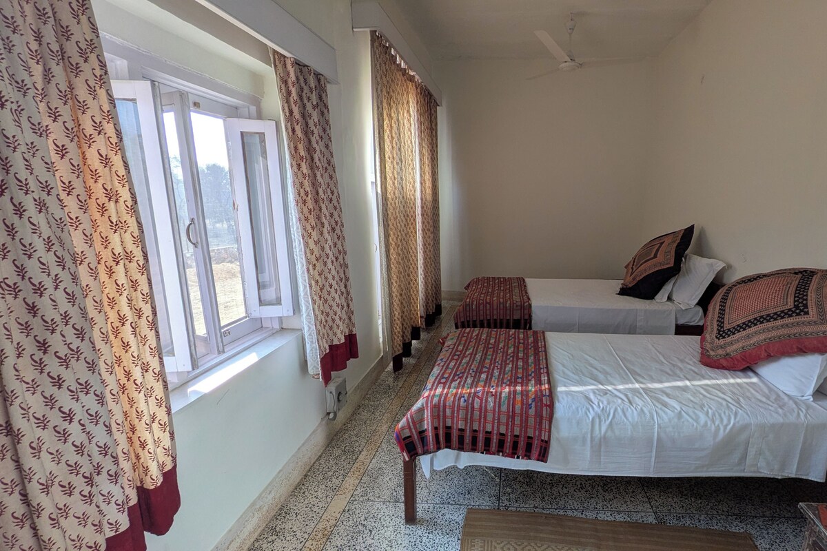A room with two beds, a window with curtains, a ceiling fan, and a patterned rug.