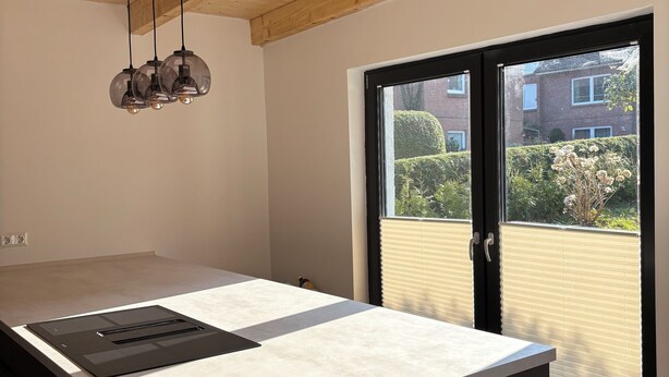 A modern kitchen with a large island, wooden ceiling beams, and a view of a residential area through a sliding glass door.