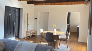 A modern living room with a grey sofa, wooden beams, a dining table with chairs, and a kitchen in the background.
