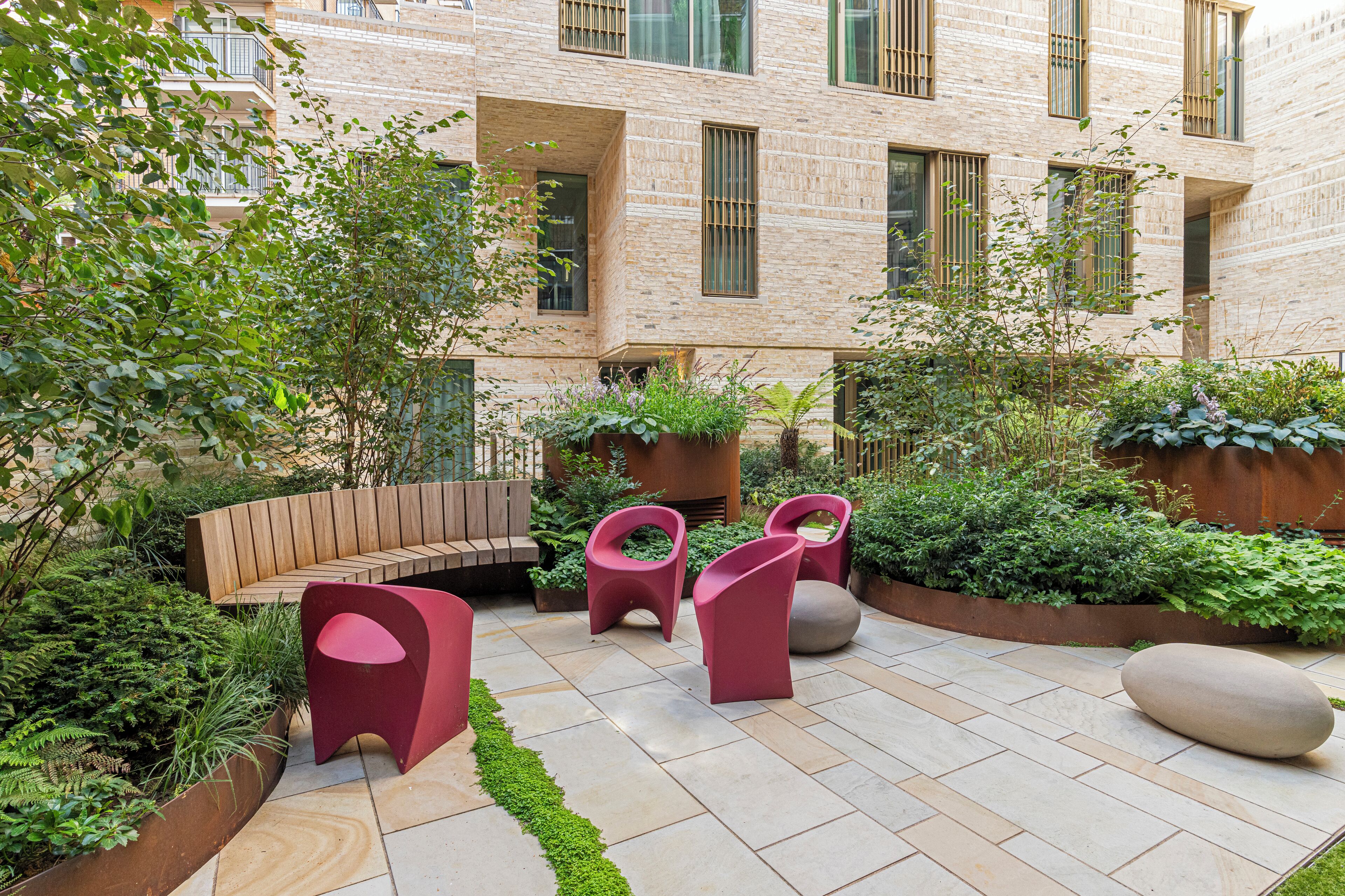 A courtyard with a wooden bench, two red chairs, and a stone planter.