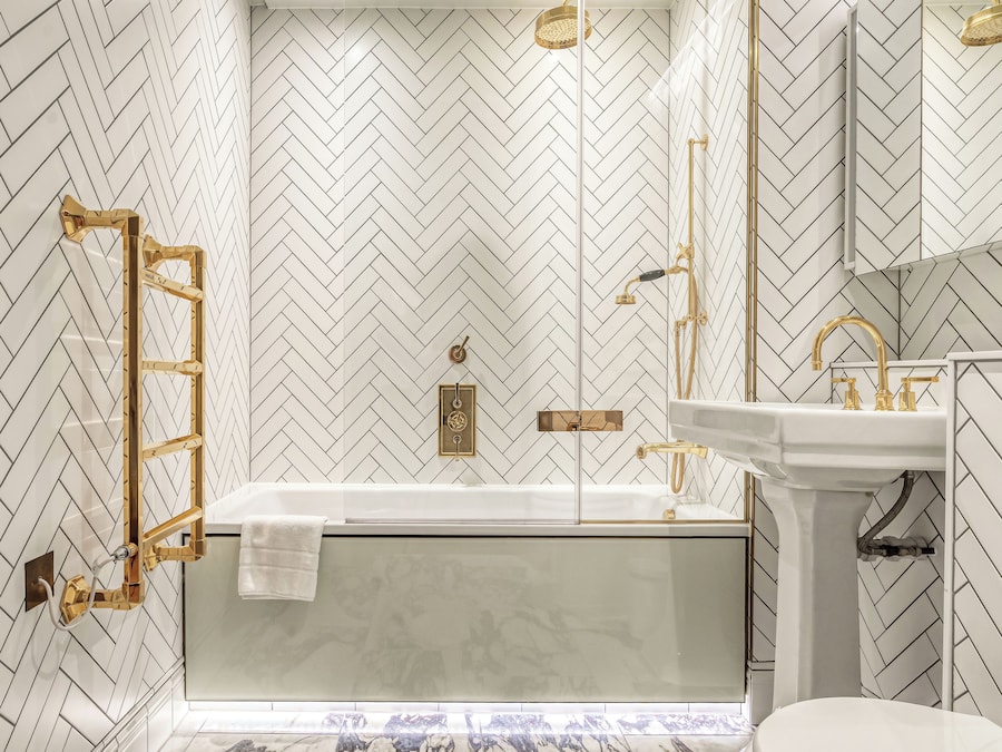 A modern bathroom with a herringbone patterned wall, a freestanding bathtub, a gold-framed towel rack, and a gold faucet.