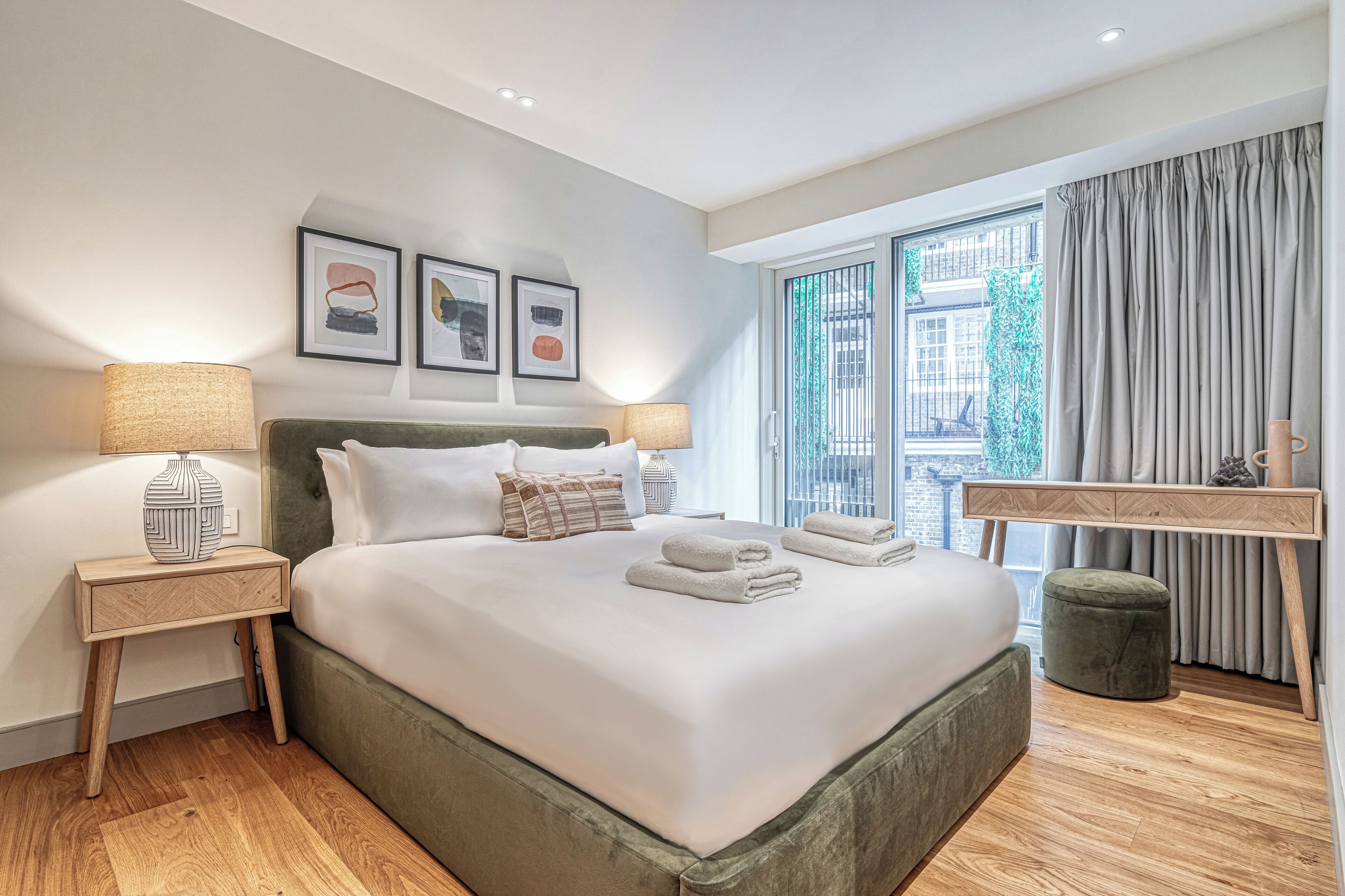 A modern hotel room with a large bed, two bedside tables with lamps, a wooden desk, and a view of the city through large windows.