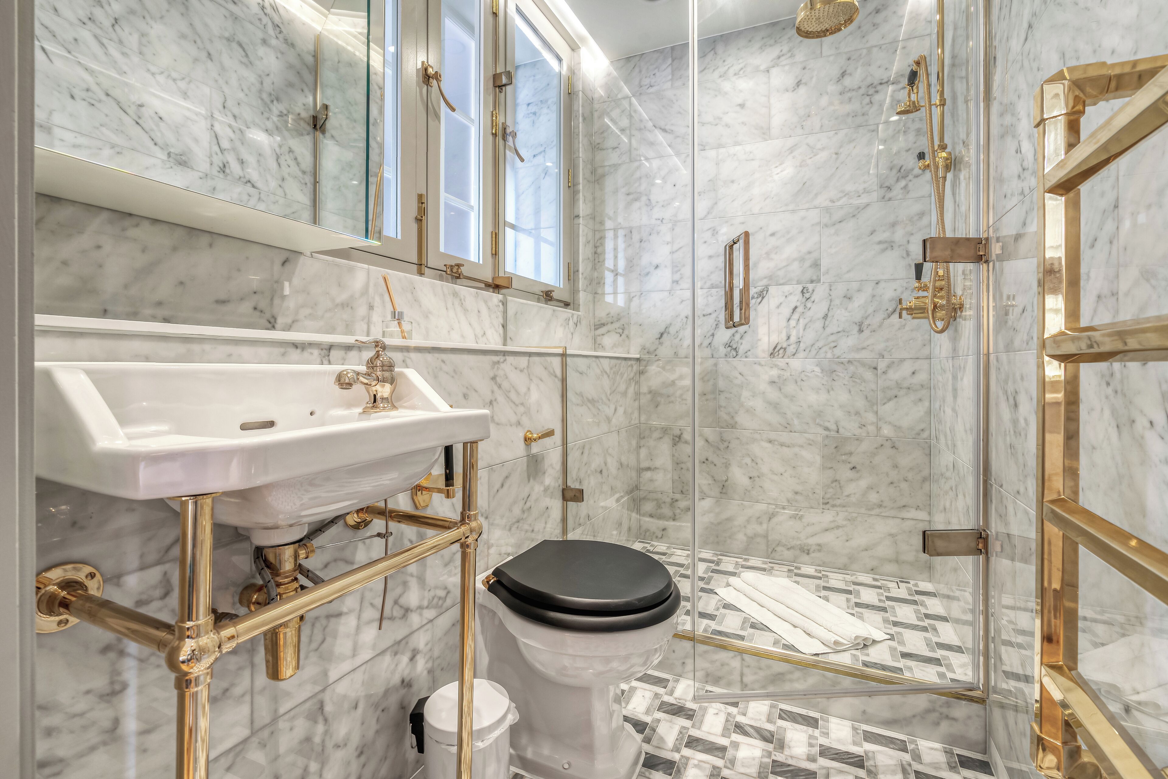 Un baño con piso de mármol, lavamanos blanco con detalles dorados, inodoro negro y zona de ducha con mampara de vidrio.
