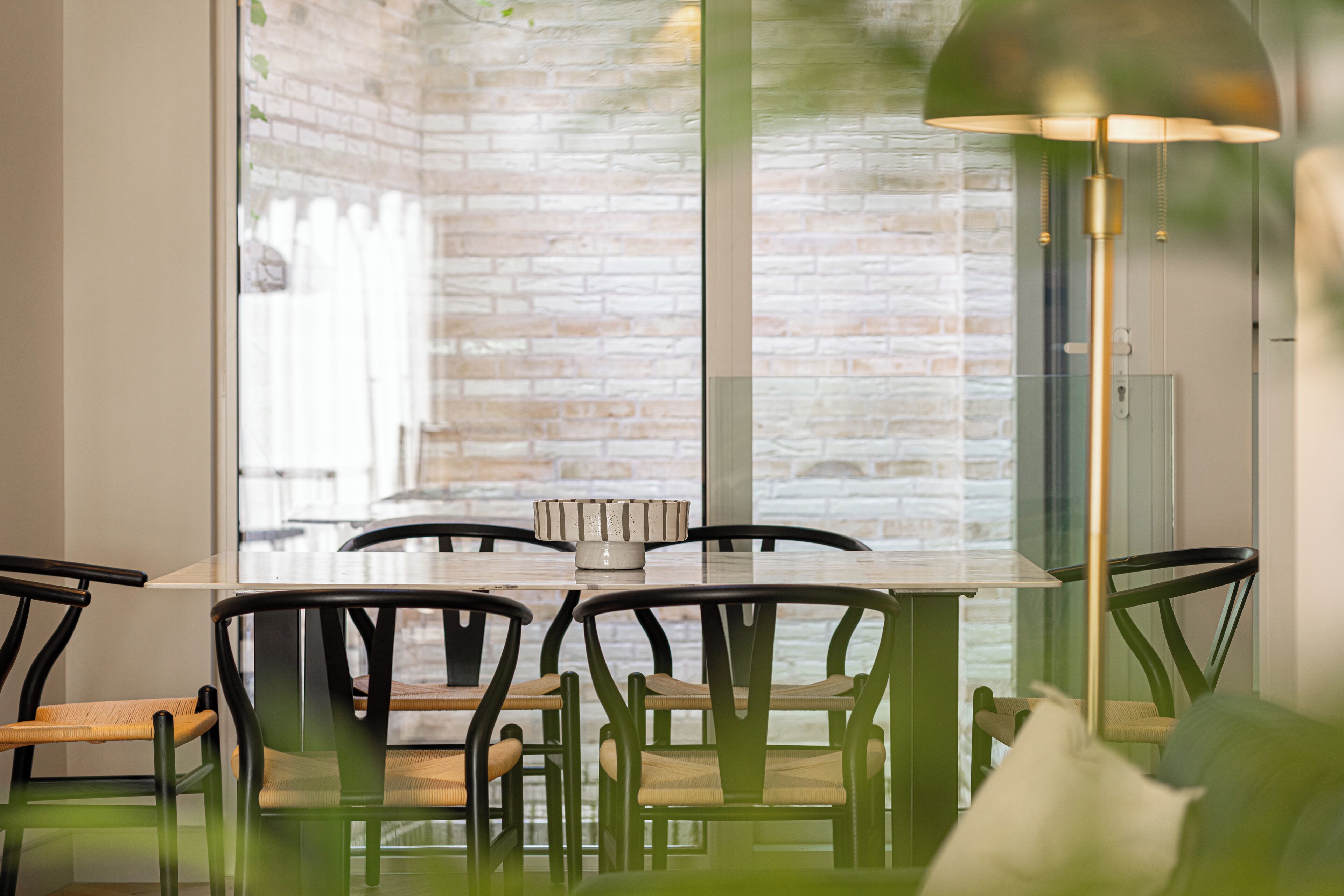 A dining area with a brick wall, a glass table, and chairs with woven seats.