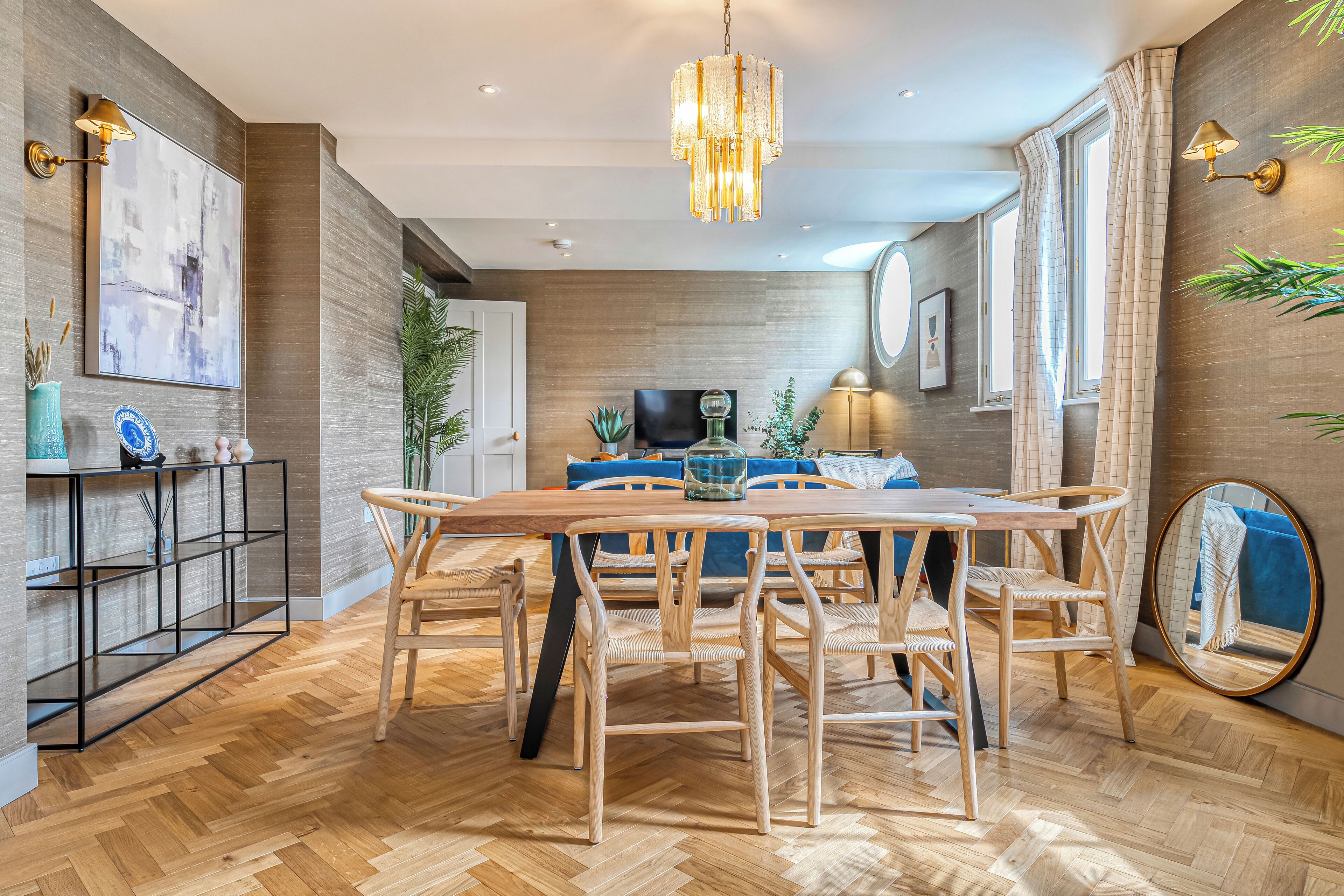 A modern dining area with a wooden table and chairs, a chandelier, and a wall-mounted lamp.