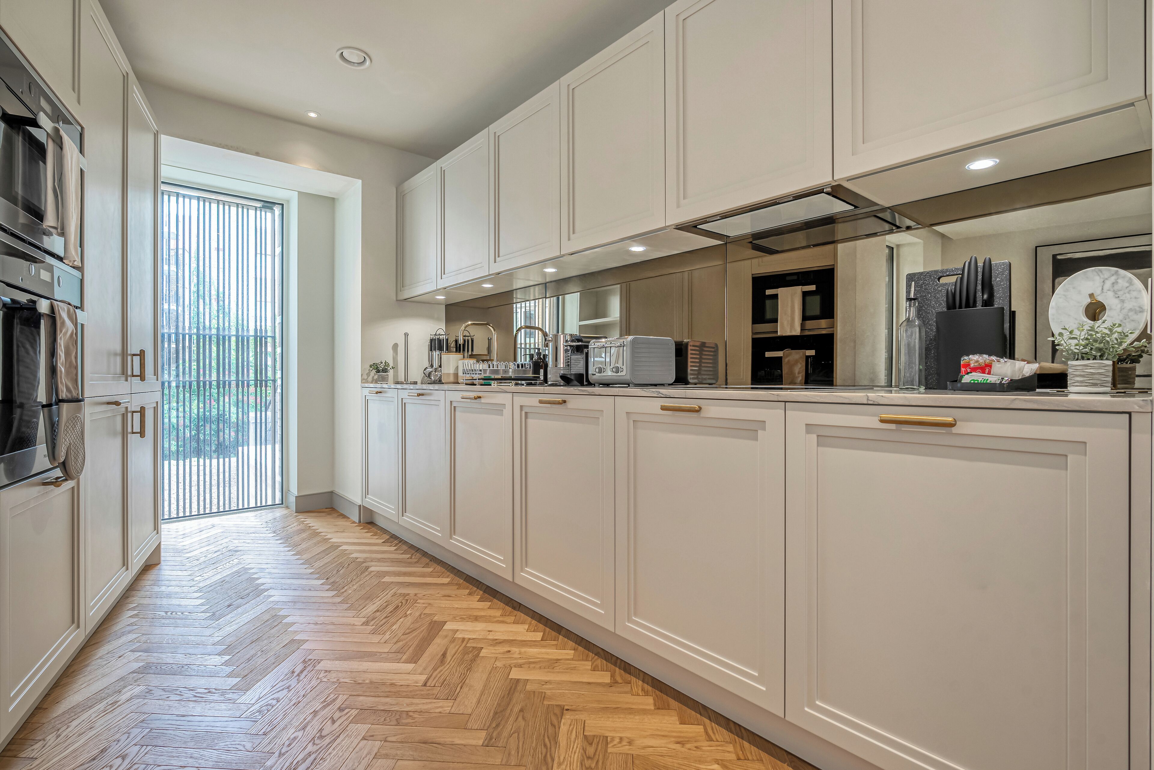 Una cocina moderna con armarios blancos, piso de madera en patrón de espiga y vistas al exterior.