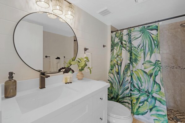 Une salle de bain moderne avec un meuble-lavabo blanc, un grand miroir et un rideau de douche orné d’un motif de feuilles tropicales.