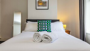 A neatly made bed with white linens and rolled towels, a checkered pillow, a bedside lamp, and a framed picture on the wall. - Beeston Central House - near shops and University (Beeston)