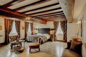 A spacious bedroom with a large bed, a sitting area with a chair and a small table, and a built-in wooden ceiling.