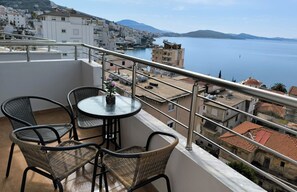 Ein Balkon mit Korbstühlen und einem kleinen Tisch, mit Blick auf eine Küstenstadt mit Gebäuden und einem Gewässer.