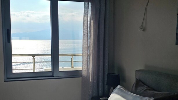 A hotel room with a bed, a nightstand, a lamp, and a window with a view of the sea and a bridge. - Albania Dream Holidays Apartment (Sarandë)