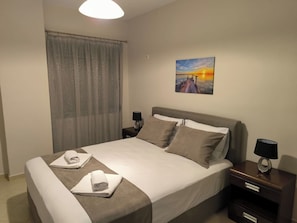 Ein ordentlich bezogenes Bett mit einer weiß-braunen Tagesdecke, zwei Kissen und zwei aufgerollten Handtüchern. Darüber an der Wand hängt ein Gemälde, das einen Sonnenuntergang zeigt.