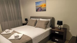Ein ordentlich bezogenes Bett mit einer weißen und braunen Tagesdecke, zwei Kissen und zwei gefaltete Handtücher an der Seite. Darüber an der Wand hängt ein Gemälde eines Sonnenuntergangs.