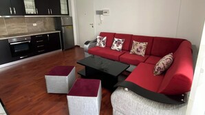 A modern living room with a red sofa, two matching ottomans, a black coffee table, and a kitchen area with dark cabinets and a white door. - Albania Dream Holidays Apartment (Sarandë)