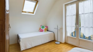 Une chambre à coucher avec un lit, une petite table, un coussin coloré, une fenêtre et une porte.