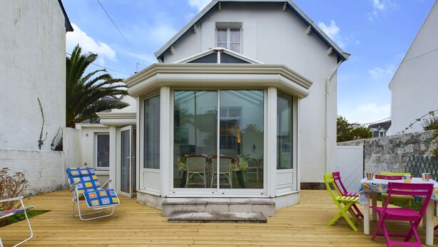 Une maison dotée d’une grande baie vitrée, d’une terrasse en bois et de mobilier d’extérieur aux couleurs vives.