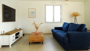 A living room with a blue sofa, a wooden coffee table with a vase, a white TV stand, and a large window. - House 6 with heated pool in St-Hilaire (Saint-Hilaire-de-Riez)