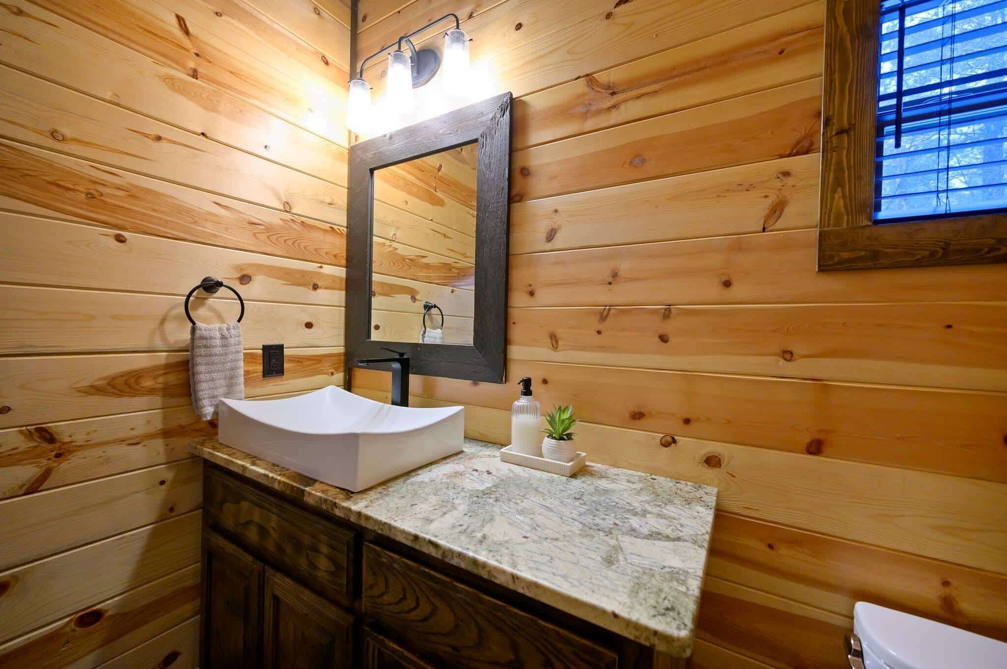 A wooden cabin bathroom with a white sink, a mirror, and a window with blinds.