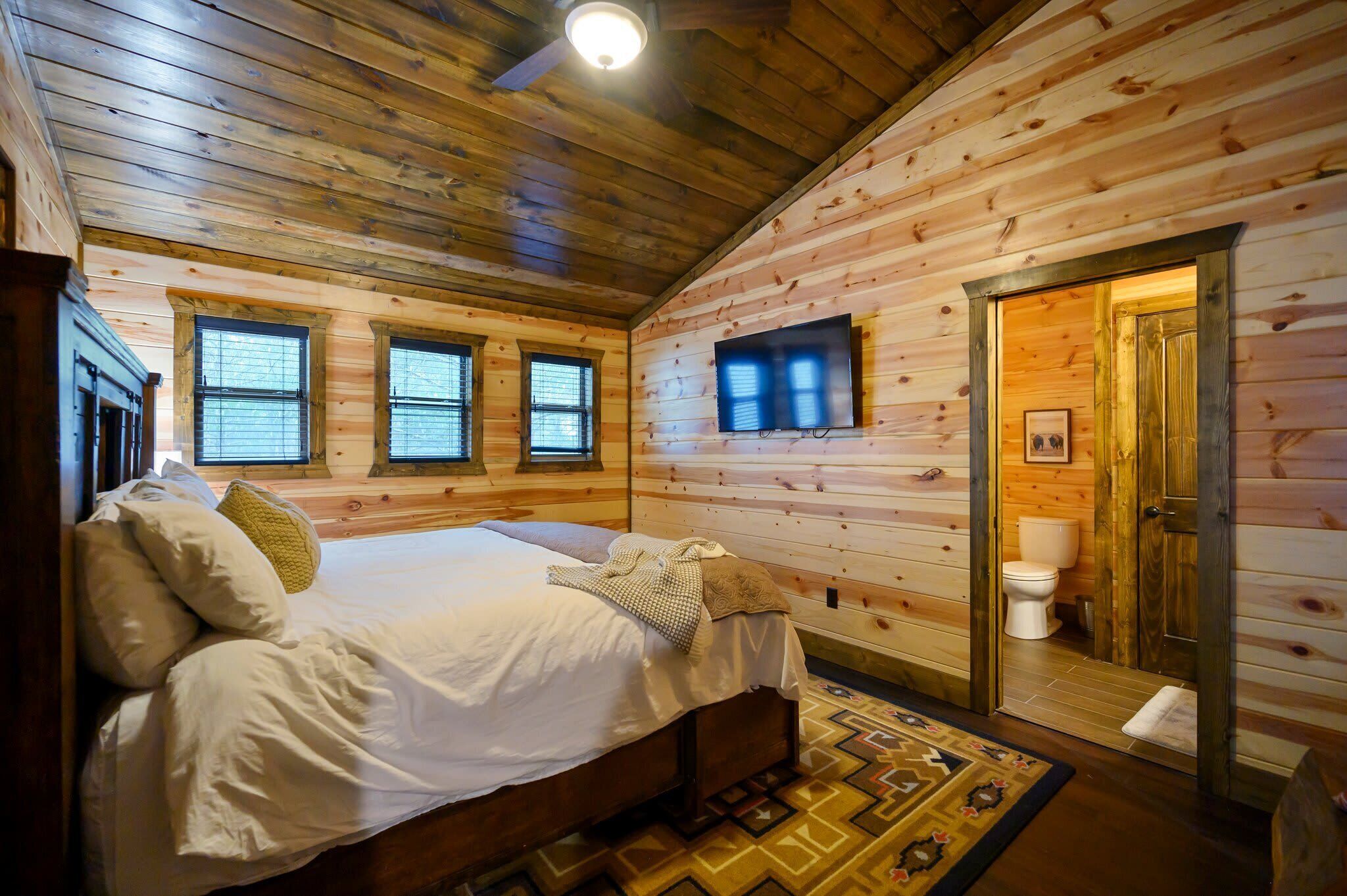 A wooden cabin bedroom with a bed, pillows, a television, and a bathroom with a toilet.