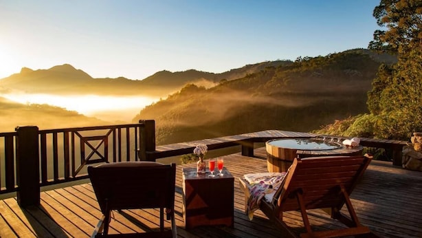 A wooden deck with a hot tub, lounge chairs, and a table with drinks, overlooking a mountainous landscape at sunrise. - Pousada Mauá Brasil (Resende)