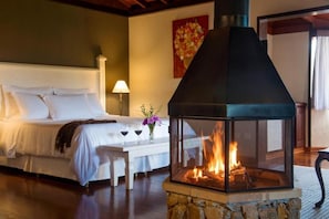 A bedroom with a stone fireplace, a bed with white bedding, a nightstand with a vase of flowers, and a painting on the wall. - Pousada Mauá Brasil (Resende)