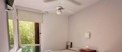 A hotel room with two beds, a ceiling fan, a small wooden nightstand, and a window with a view of greenery.
