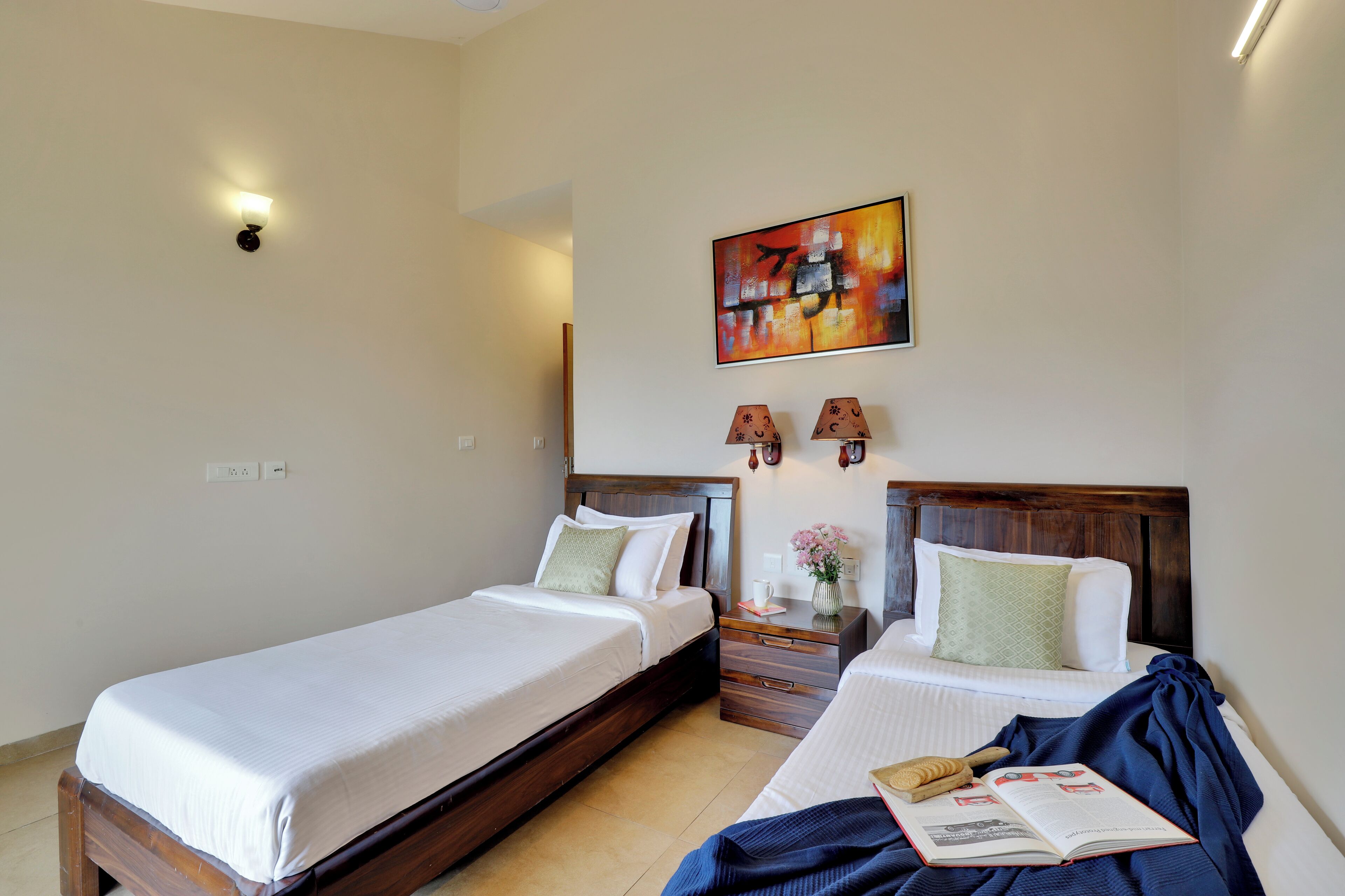A hotel room with two single beds, a nightstand with a lamp, a painting on the wall, and a book on the bed.