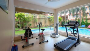 Une salle de sport à domicile équipée de matériel d’exercice, avec un ventilateur de plafond et une vue sur une piscine et la verdure environnante.