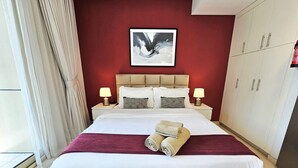 Una habitación de hotel con una cama grande, dos lámparas de noche, una pared de acento roja y una obra enmarcada sobre la cama.