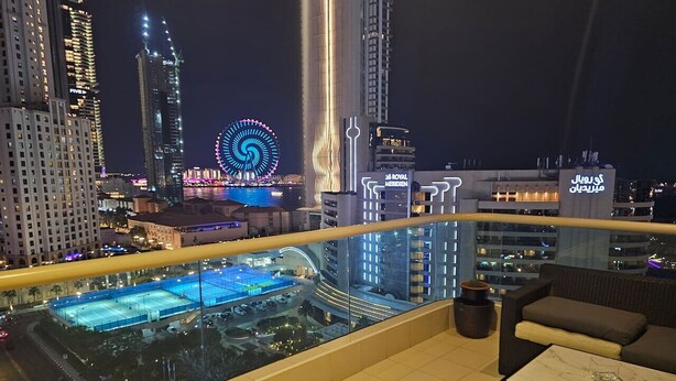A high-rise hotel room with a balcony overlooking a cityscape, including a Ferris wheel and modern buildings. - Sunrise Homes - Studio with Beautiful View next to JBR Walk and Beach (Dubai Marina)