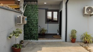 Een inkomst van een woning met een betegeld terras, planten in potten, een airconditioning en een raam met uitzicht.