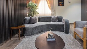 Een moderne woonkamer met een hoekbank, een ronde salontafel, een houten bijzettafeltje, een potplant en een geometrische plafondlamp.