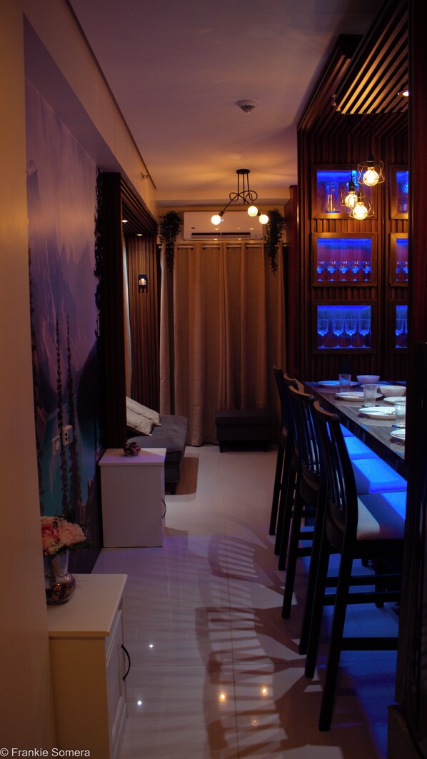 A dimly lit room with a dining area featuring a long table, chairs, and a bar area with shelves.