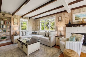 A cozy living room with a sectional sofa, a coffee table, a striped armchair, a fireplace, a bookshelf, and a window with a view of greenery.