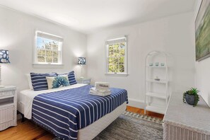 Un dormitorio con cama, dos ventanas, una mesita de noche, lámpara, estante y planta en maceta.