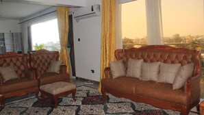 Ein Wohnzimmer mit zwei braunen Sofas und einem passenden Hocker, einem gemusterten Teppich und Blick auf eine Stadtlandschaft durch große Fenster.