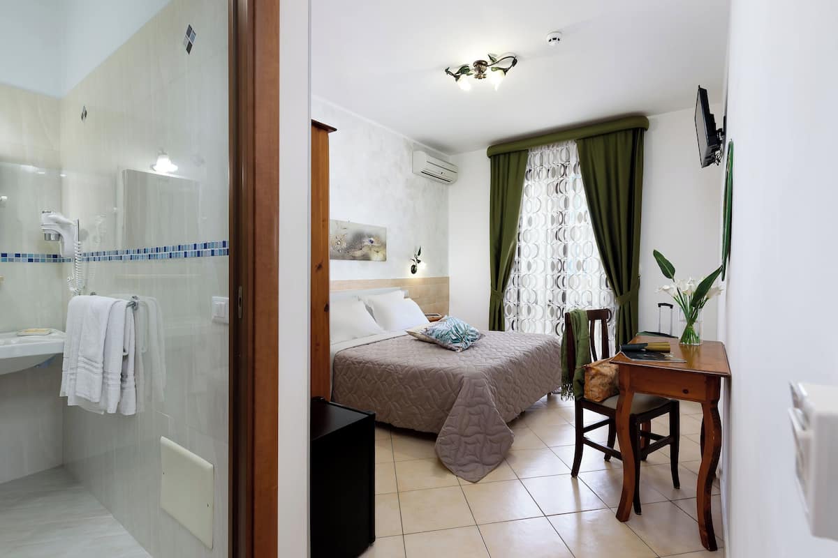 A bedroom with a bed, a bedside table, a chair, a desk, a mirror, and a shower area.