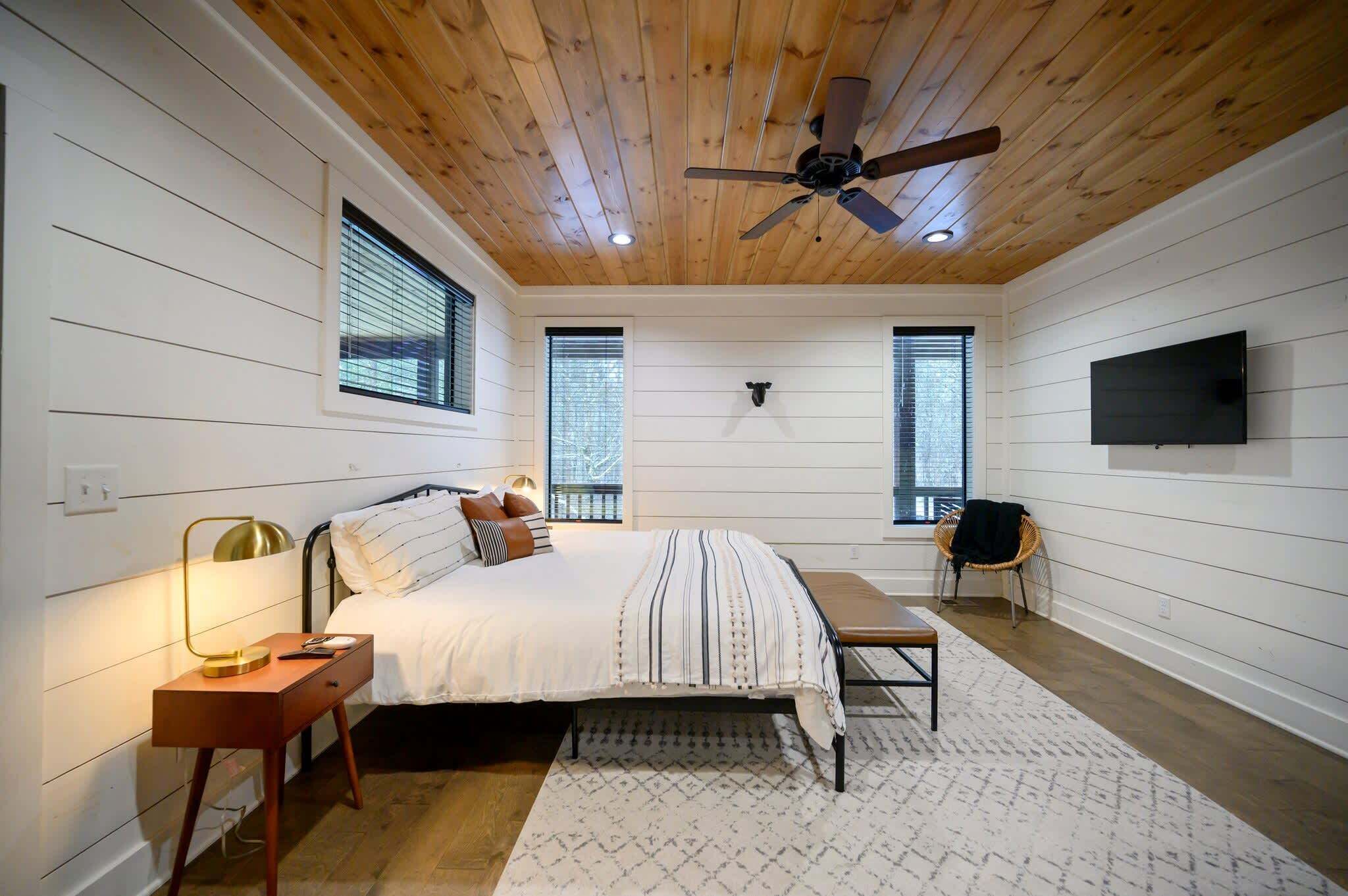 A bedroom with a wooden ceiling, a bed with a striped bedspread, a bedside table with a lamp, and a TV mounted on the wall.