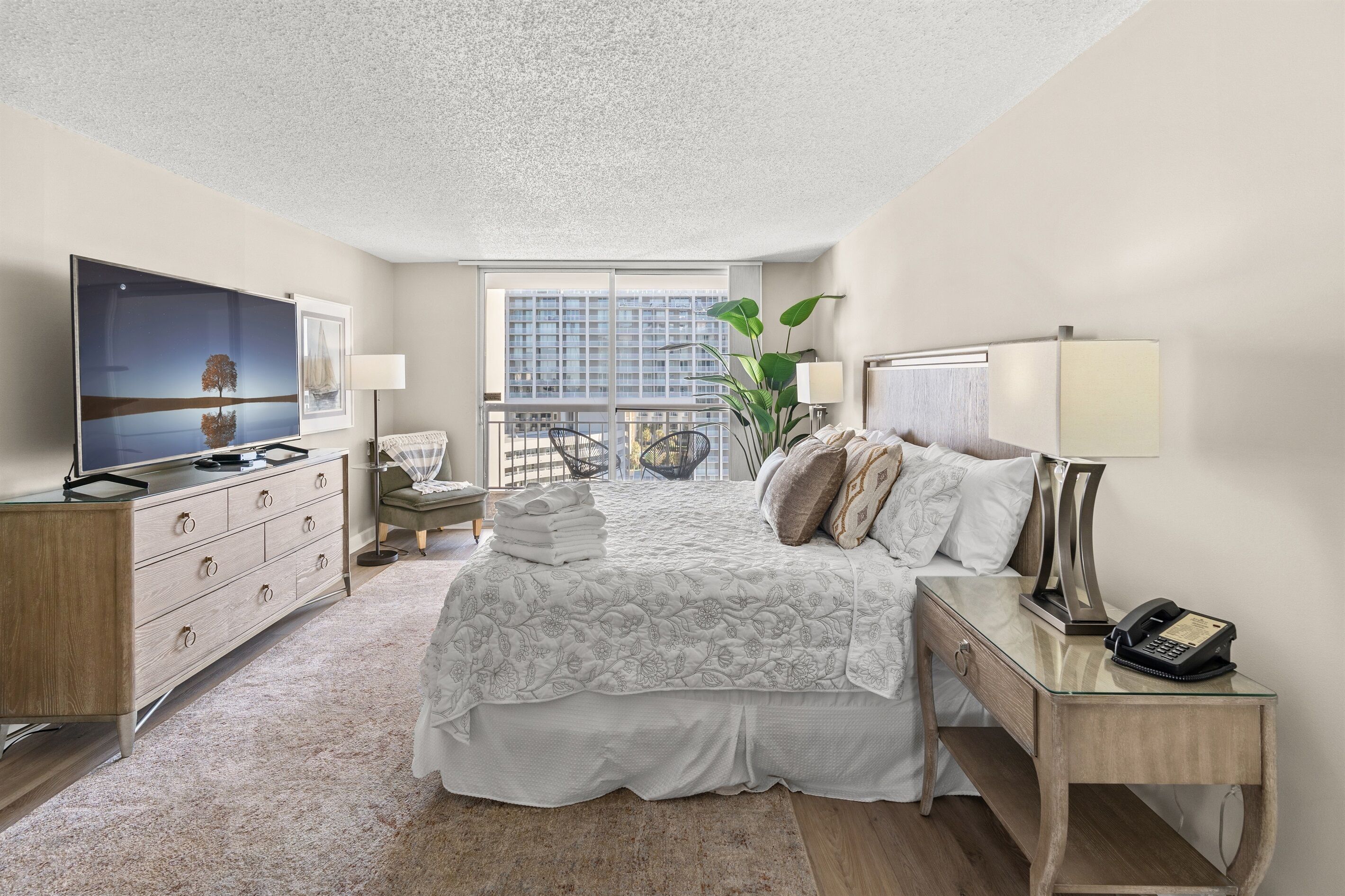 A hotel room with a large bed, a TV, a desk with a lamp, and a view of the city through large windows.