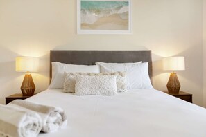 A neatly made bed with white linens and two decorative pillows, flanked by two bedside tables with lamps, in a room with a simple, clean design. - Palm View- 9 Pools BBQ Gym (Westcourt)