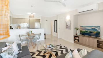 Una sala de estar moderna con sofá, mesa de centro y televisor. Cuenta con una zona de comedor con mesa y sillas. La habitación tiene ventilador de techo y aire acondicionado.
