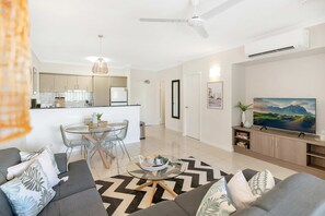 A modern living room with a sofa, coffee table, and a TV. There is a dining area with a table and chairs. The room has a ceiling fan and an air conditioning unit. - Palm View- 9 Pools BBQ Gym (Westcourt)