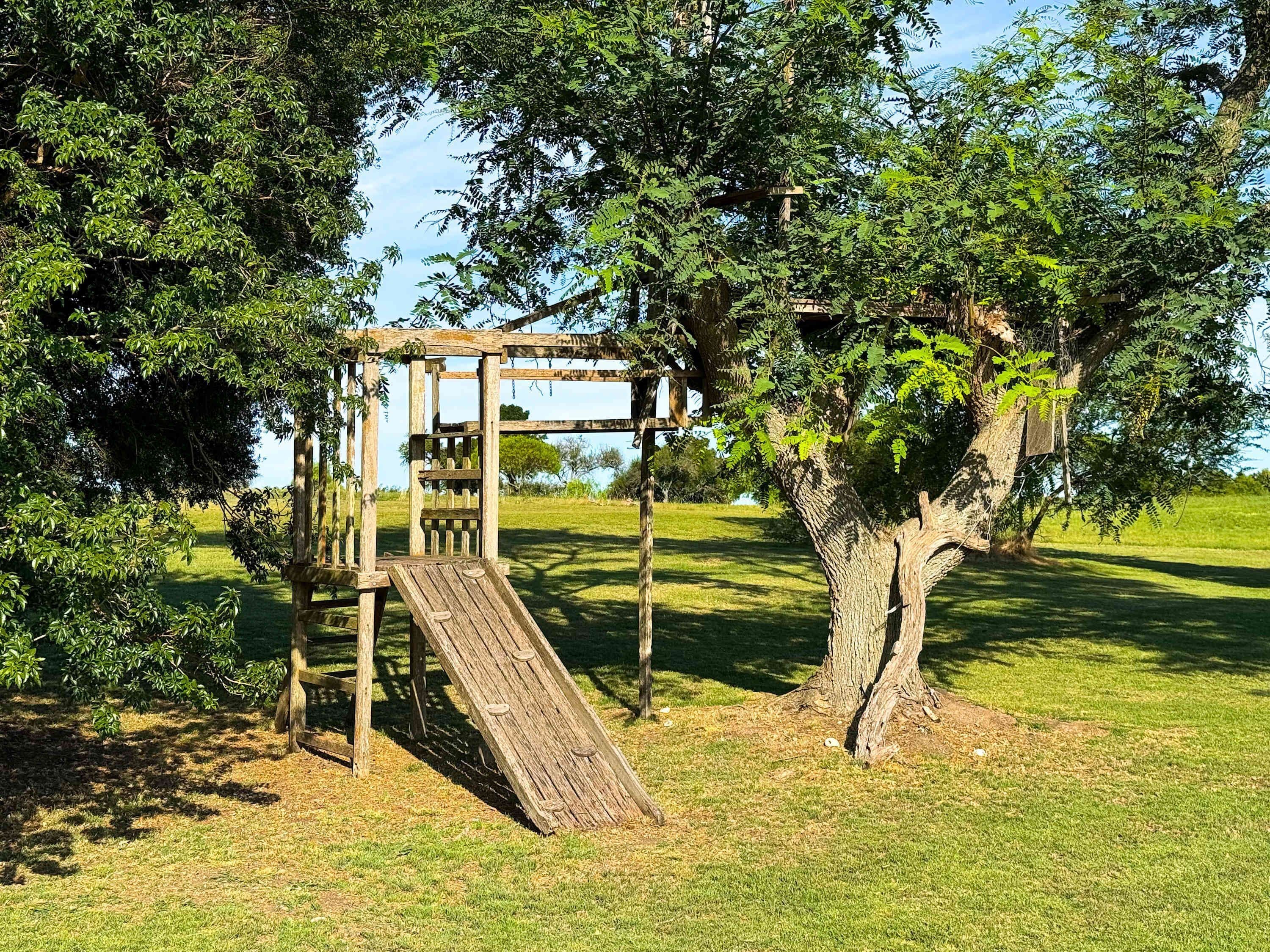 Estructura de juegos de madera con un tobogán, rodeada de árboles y césped.